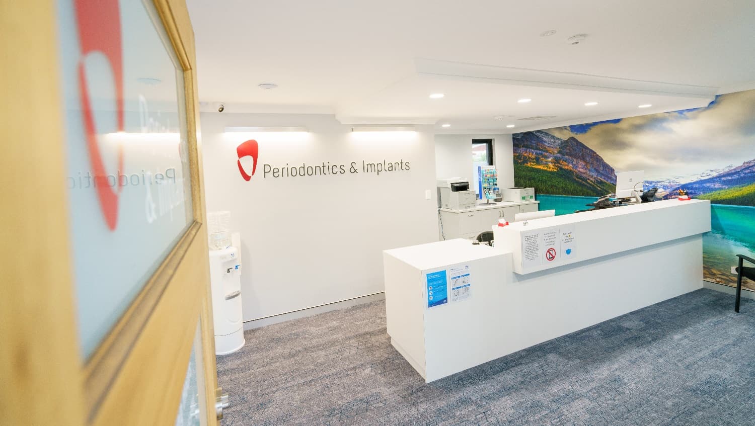Upon entry to Periodontics & Implants front door, you will see a white-water dispenser that connects to the white wall and ceiling. The whole floor is carpeted and the long reception desk is painted white. A large wall further to the right has a landscape picture of clouds, mountains, and water that adds a soothing element to the room. A white cabinet is placed at the back corner of the long desk while securing a space between them, to give more space for the clinic personnel.