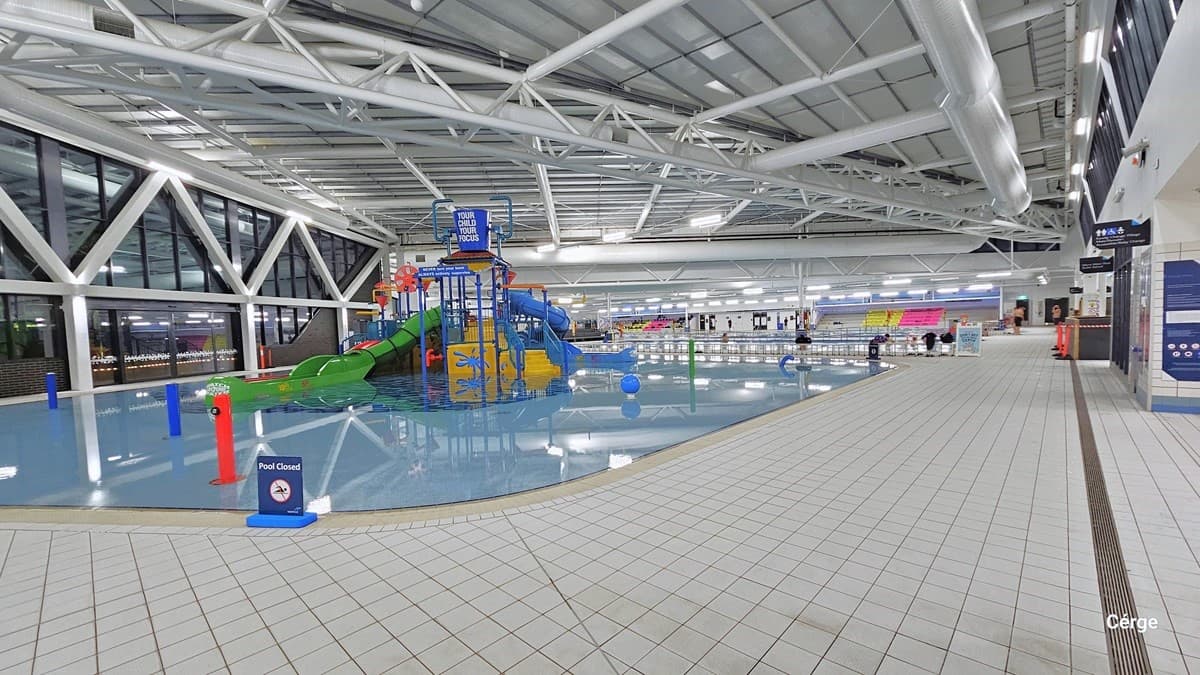 Here there is a shallow beach entry pool with two enclosed tube slides. The pool is equipped with interactive water cannons and a blue tipping bucket positioned overhead. The prominent white ceiling shows white pipes and tubes. There's a freestanding blue warning sign that reads "Pool Closed." The floor is tiled in white.  On the left, a glass wall features a large steel "W" design suspended from the ceiling. To the right, banners are on display, providing instructions and directions.