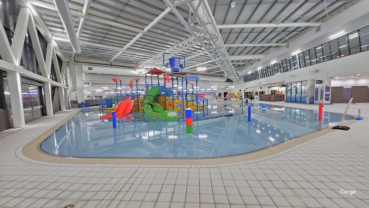 Another view of the shallow beach entry pool with two enclosed tube slides. The pool is equipped with interactive water cannons and a blue tipping bucket positioned overhead. The prominent white ceiling shows white pipes and tubes. There's a freestanding warning sign on the right. The floor is tiled in white.  On the left, a glass wall features a large steel "W" design suspended from the ceiling. To the right, banners are on display, providing instructions and directions.