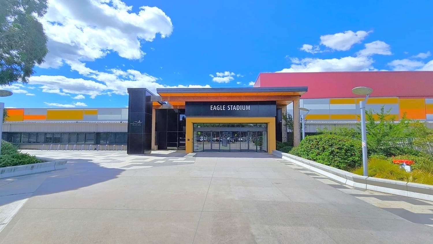 Eagle Stadium facility with a distinctive wall color scheme of yellow, white, and red rectangular tiles. The building has a unique build, with its end looking like it is sloping downward. The triangular front is made of glass walls with black metal frames. There is a large orange arrow pointing straight ahead on the upper right glass wall. Eagle Stadium is detailed in large white letters above the glass wall on the left glass wall. Adjacent to the facility are gardens and tall trees.