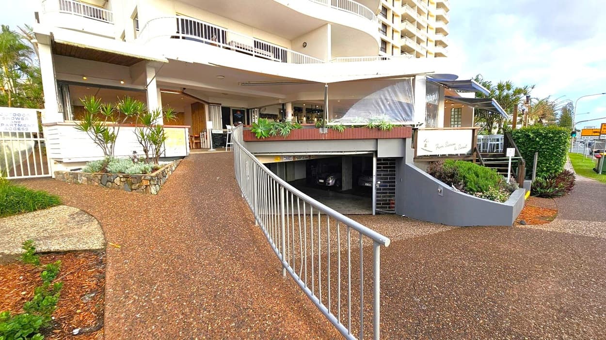 A building with a curved design features a ramp leading to an underground car park on the right. Above the car park entrance, there is a small garden with potted plants. To the left, a stone planter with greenery is adjacent to a sign indicating outdoor shower and apartment access. The building has balconies with white railings. A pathway with a textured surface runs alongside the structure, and there are tables and chairs visible near the entrance.