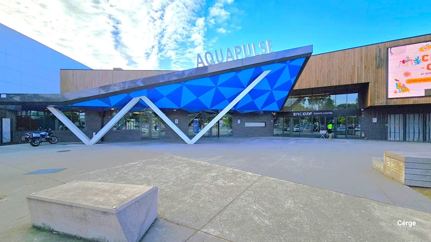 Aquapulse is housed within a substantial building. To the left of the entrance, you'll find a large, winding water slide with blue and green hues. The entrance is encircled by landscaped gardens and trees. The automatic glass sliding doors grant access to Aquapulse. Just before the entrance, you'll notice a distinctive "W" design and a ceiling in blue and green with triangular patterns.