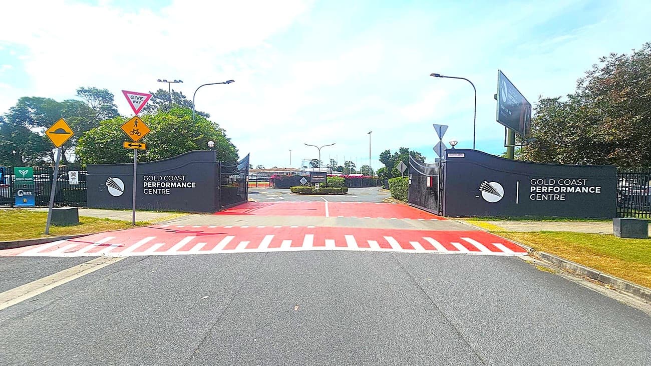 Entrance to the Gold Coast Performance Centre with black gates on both sides. Signs on the left include a "Give Way" and a pedestrian/cyclist warning. The ground features a red and white striped pedestrian crossing. Trees and bushes are visible in the background, along with a few streetlights. The road is grey, and the area is well-maintained.