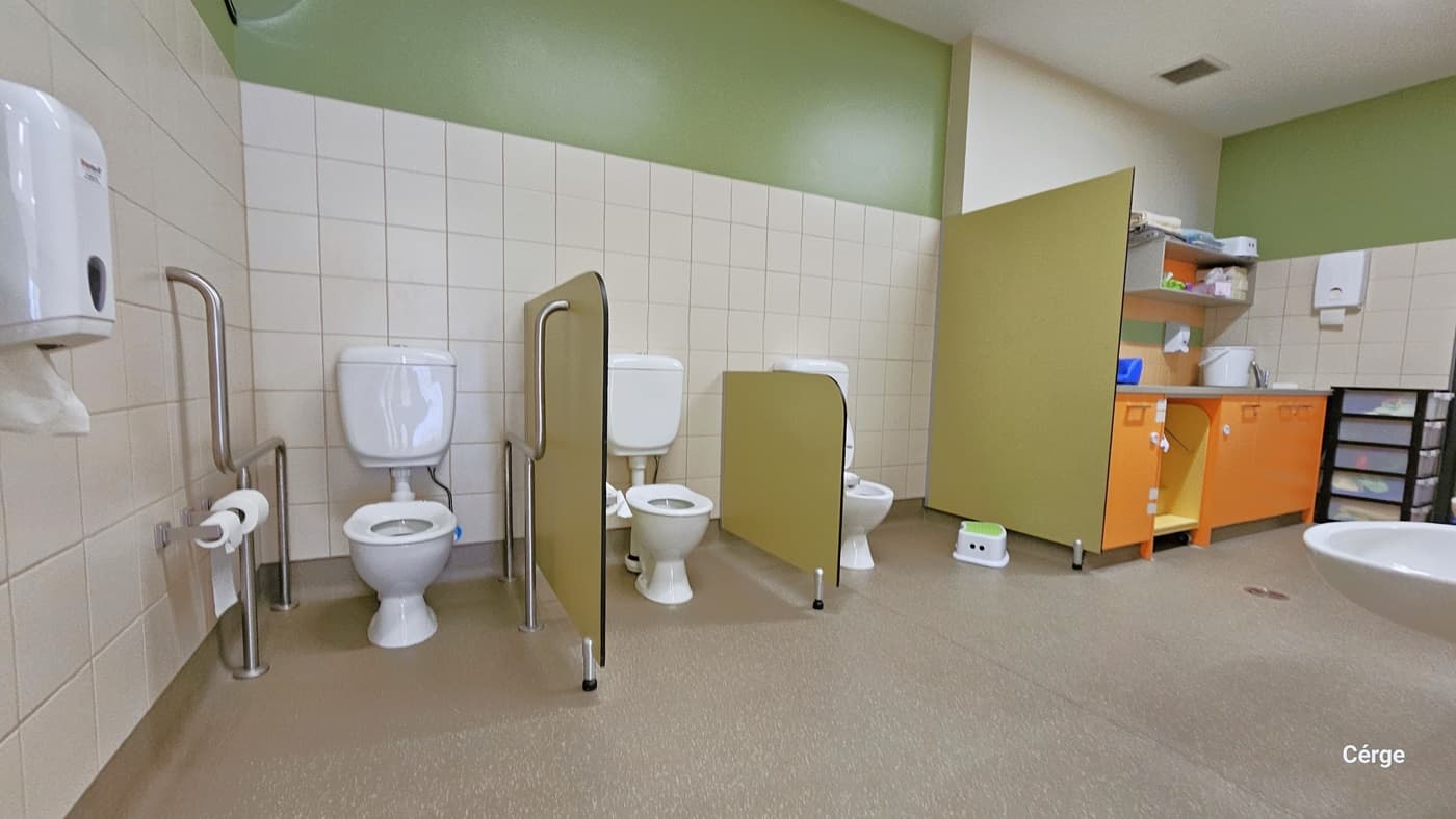 Inside the bathroom of Crèche at Eagle Stadium. The bathroom has a natural grey stone tile floor and a white tiled wall on the lower portion, while the upper part is green. Each toilet is separated by a small lime green wall cover supported by metal frames. The toilet near the left wall has grab bars on both sides and has a roll of tissues on them. There is also a liquid hand soap dispenser, a wash basin, and other toilet kits and accessories on the orange cabinet on the far right.