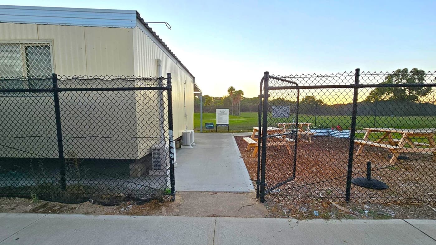 A chain-link fence with an open gate is in the foreground, leading to a concrete path. On the left, a light-coloured portable building with a barred window and an air conditioning unit is visible. To the right, there are wooden picnic tables on a dirt area. In the background, a grassy field with trees and a sign can be seen. The sky is light, suggesting early morning or late afternoon. The ground is a mix of concrete and dirt.