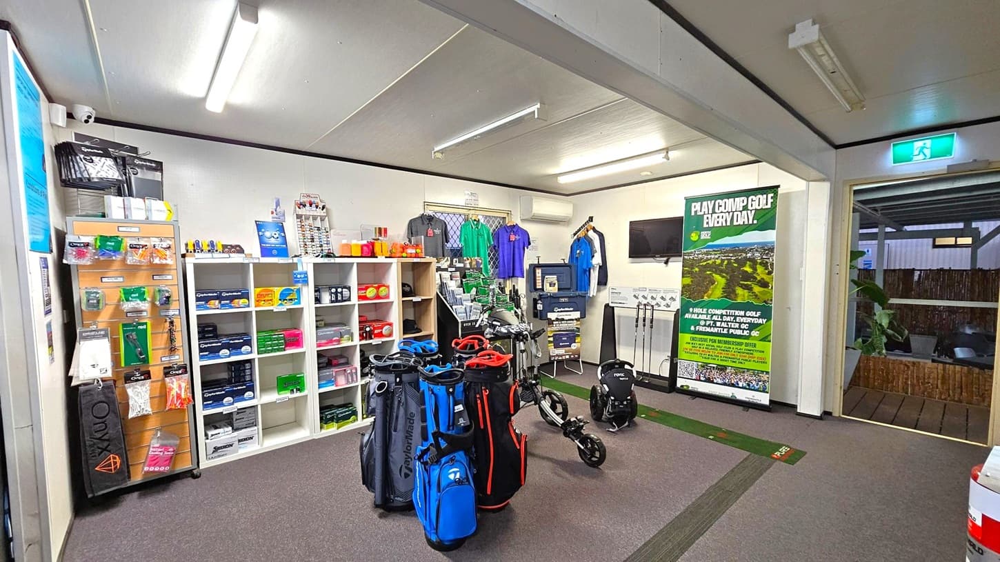 A golf shop interior with shelves on the left displaying various golf accessories like balls, tees, and gloves. In the centre, several golf bags in blue, black, and red are on display alongside a golf trolley. The back wall features a selection of polo shirts in different colours. A large poster promoting golf competitions is on the right, near a small TV mounted on the wall. The floor is carpeted, and the ceiling has fluorescent lighting. An exit sign is visible above a doorway on the right.