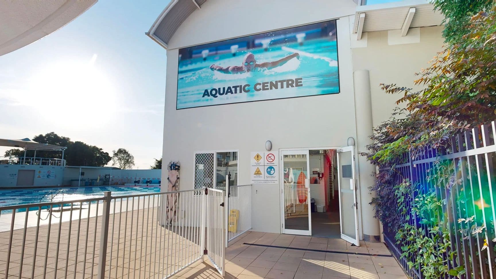 On the left, a swimming pool surrounded by a low metal fence and a shaded seating area in the background. In the middle, a white building with an open glass door leading to the interior, a caution sign near the entrance, and a large poster above featuring a swimmer with the text "Aquatic Centre". On the right, a tall black metal fence with green foliage alongside. The tiled floor in the foreground is light beige, and the building has a curved metal roof overhang.