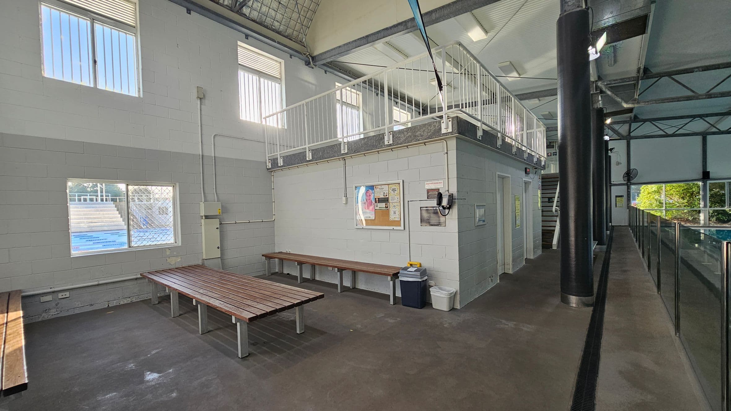 On the left, wooden benches sit against a grey cinder block wall with three windows, one of which overlooks an outdoor pool. In the middle, a raised area with white railings supported by a boxy structure holds a noticeboard, two bins, and a wall-mounted telephone. On the right, black columns and a glass partition run alongside an indoor swimming pool. The floor is dark concrete, and the ceiling includes exposed beams, metal trusses, and white panels with fluorescent strip lights.