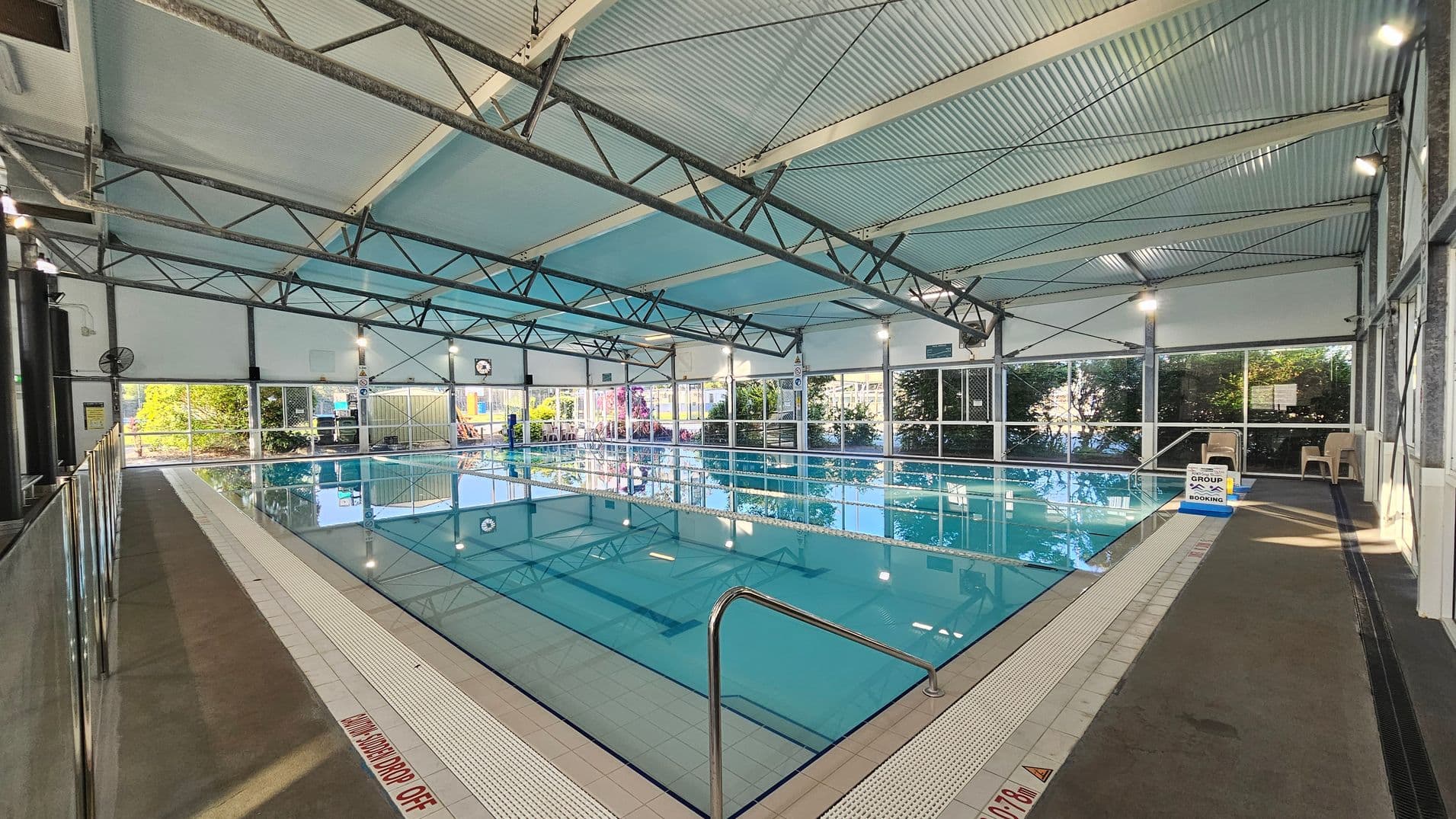 On the left, a grey concrete walkway with a stainless steel barrier runs alongside the pool. In the middle, a rectangular swimming pool with clear blue water is surrounded by white tiles, featuring depth markings near the edges and a handrail in the foreground. On the right, beige plastic chairs are placed on the dark non-slip flooring next to a glass-panelled wall. The ceiling is corrugated metal with exposed beams and overhead lights, supported by metal trusses.