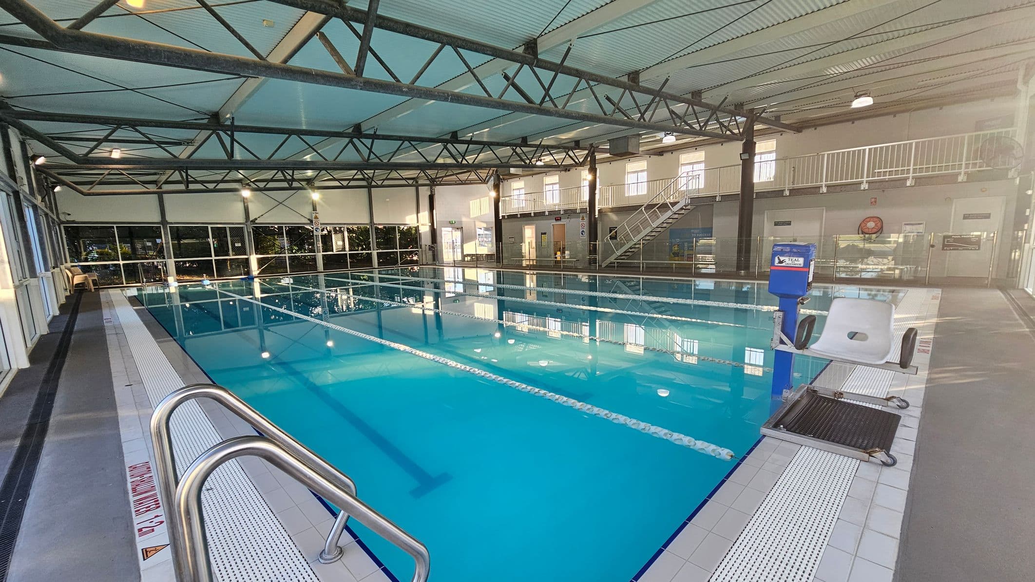 On the left, a long walkway with dark grey non-slip flooring runs beside the pool, with white chairs and glass wall panels in the background. In the middle, a rectangular swimming pool with bright blue water is divided by lane ropes, surrounded by white tiled edges. On the right, a pool chair lift is positioned over the water, with a staircase and mezzanine level visible in the background. The ceiling has corrugated metal panels, exposed beams, and suspended lights.