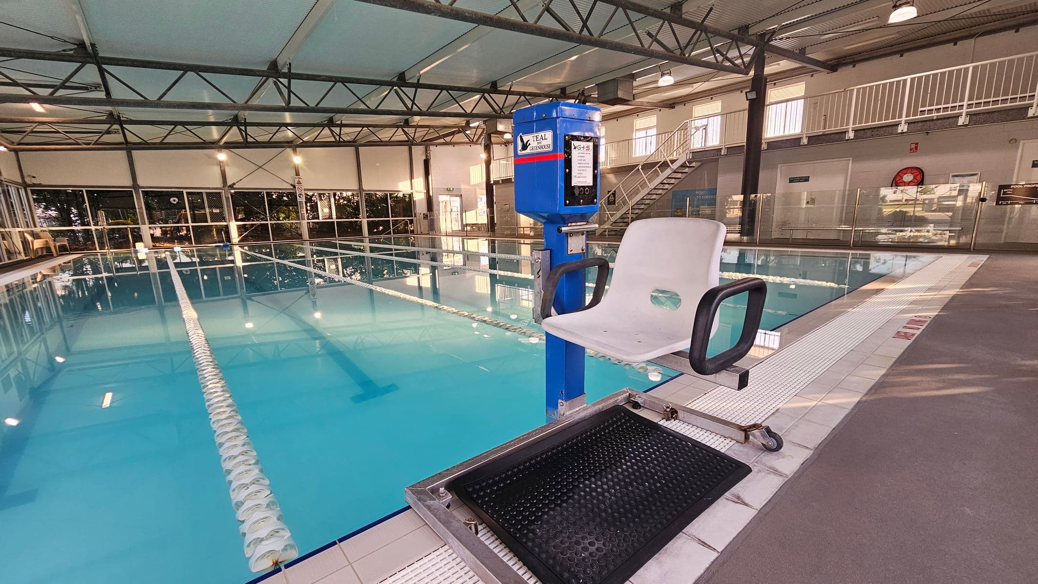 On the left, a swimming pool with clear blue water is divided by white lane ropes, with beige chairs and a glass-panelled wall in the background. In the middle, the pool extends towards a wall featuring glass doors, metal frames, and greenery outside. On the right, a pool chairlift with a black platform stands beside the pool, near glass barriers and a staircase leading to a mezzanine. The floor is tiled around the pool and grey elsewhere.