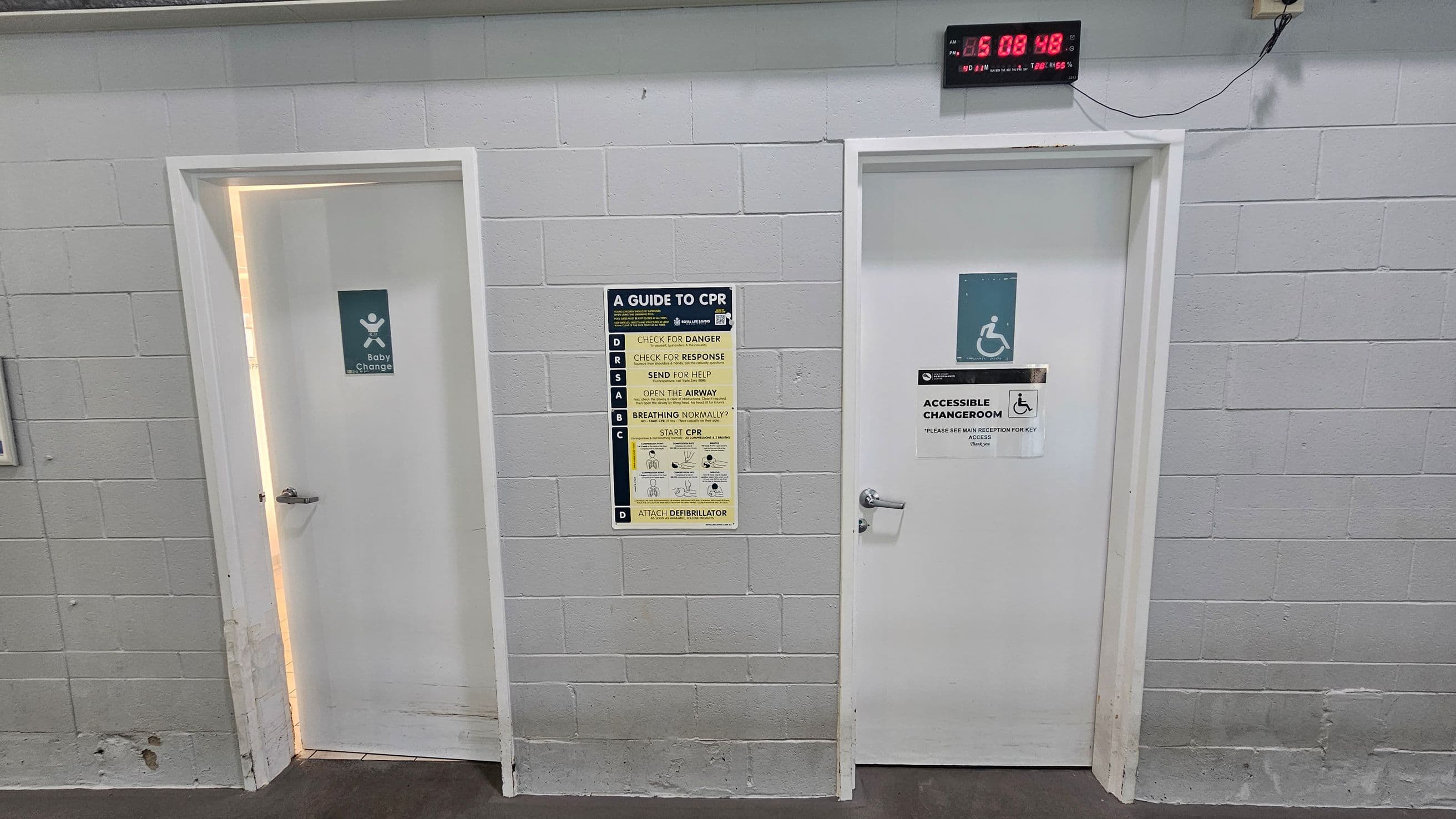 On the left, a white door slightly ajar with a baby change sign is set into a grey cinder block wall. In the middle, a CPR guide poster is mounted on the wall, listing instructions in yellow and black with a diagram. On the right, a closed white door labelled "Accessible Changeroom" is below a wall-mounted digital clock displaying red numbers. The floor is smooth grey concrete, and the ceiling features exposed metal beams and piping.
