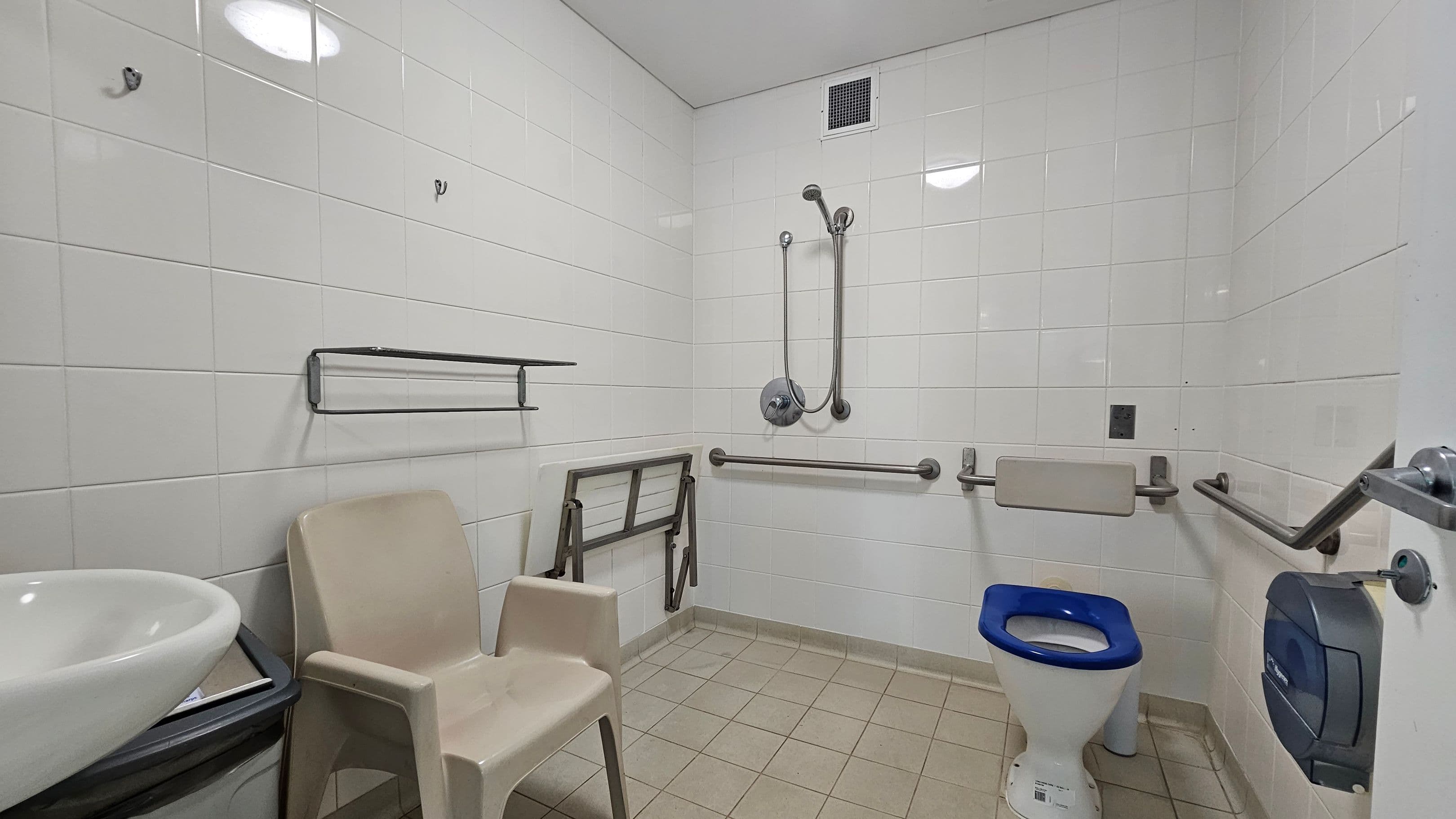 On the left, a beige plastic chair is next to a small white basin with a black edge. Above the chair, metal hooks and a towel rack are mounted on the white tiled wall. In the middle, grab rails and a foldable support bar are fixed to the wall beside a handheld showerhead. On the right, a white toilet with a blue seat is positioned below more grab rails, with a mounted toilet paper dispenser nearby.