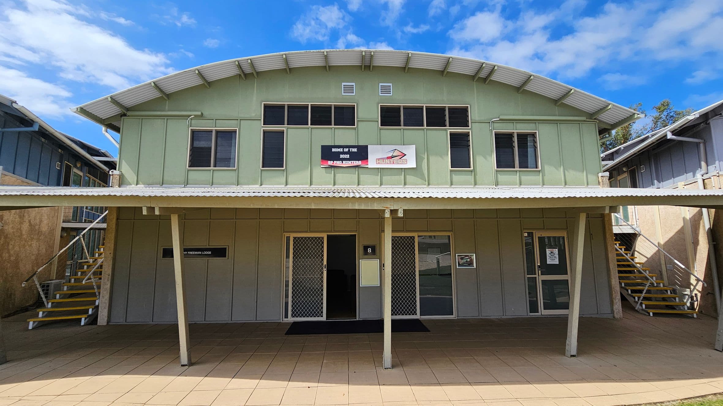 A two-storey building with a green facade and a corrugated metal roof. The upper level has several windows and a sign reading "Home of the 2022 SP PNG Hunters." The lower level features two doors with security screens and a sign for "Freeman Lodge." On both sides, there are metal staircases leading to the upper level. The ground is paved with beige tiles.