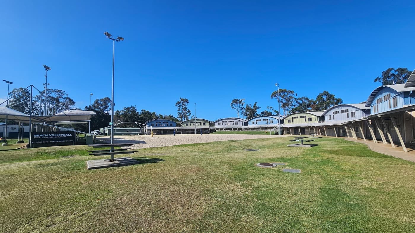 A grassy area with a beach volleyball court in the centre, surrounded by a row of curved-roof buildings in the background. On the left, there's a sign reading "Beach Volleyball Social Development Elite Gold Coast Performance Centre" near a covered seating area. Several tall floodlights are visible, and a picnic table is in the foreground. Trees are scattered in the background.