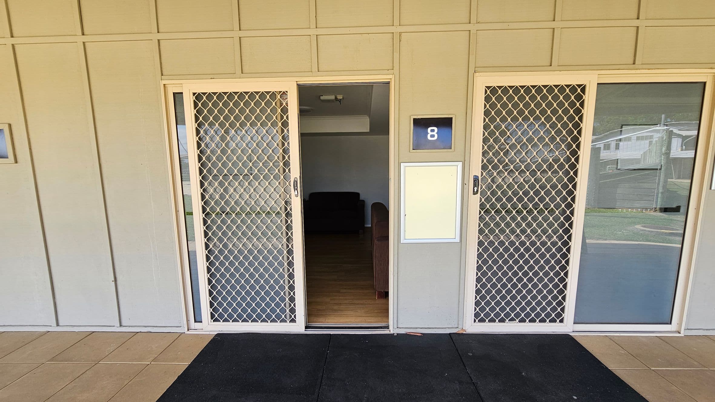 Sliding glass doors with white mesh screens are in the centre, partially open to reveal a dimly lit interior with a dark sofa. A blue sign with the number 8 is mounted on the wall to the right of the door. The exterior wall is light-coloured with vertical panels. The floor is tiled in a light beige, with a dark mat in front of the entrance. Reflections of nearby buildings are visible in the glass on the right.