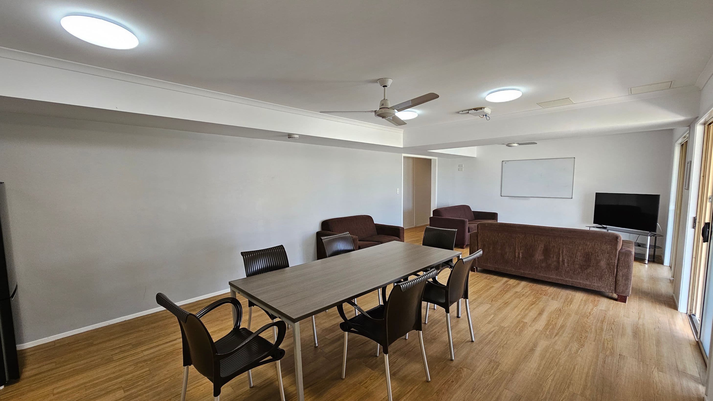 A room with wooden flooring and a white ceiling featuring recessed lights and a ceiling fan. On the left, there's a long table with six black chairs. In the background, a brown sofa and two armchairs face a flat-screen TV on a stand. A whiteboard is mounted on the wall behind the sofa. A doorway is visible in the centre, and large windows on the right allow natural light to enter.
