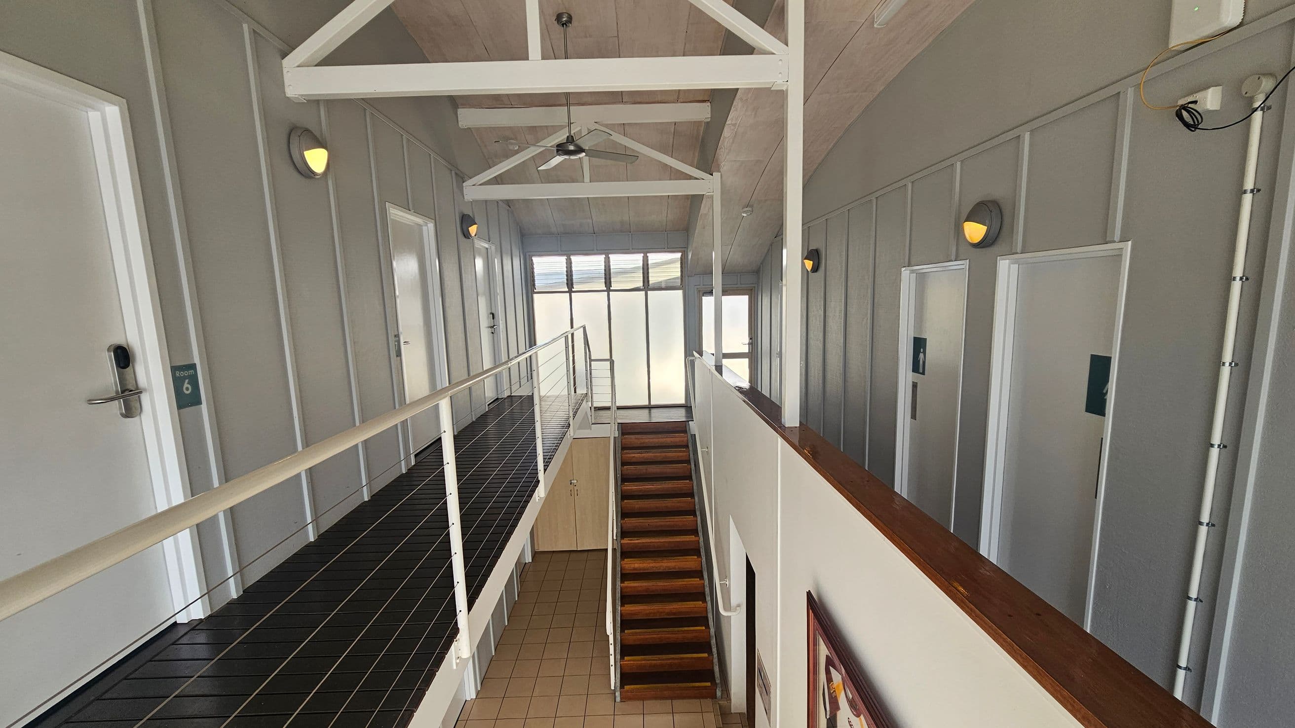 A corridor with a black metal walkway on the left, featuring white doors labelled "Room 6" and others. The right side has grey walls with round wall lights. In the centre, a wooden staircase leads down to a tiled floor. The ceiling has exposed white beams and a ceiling fan. At the far end, large windows allow natural light to enter. A framed picture is visible on the wall near the staircase.