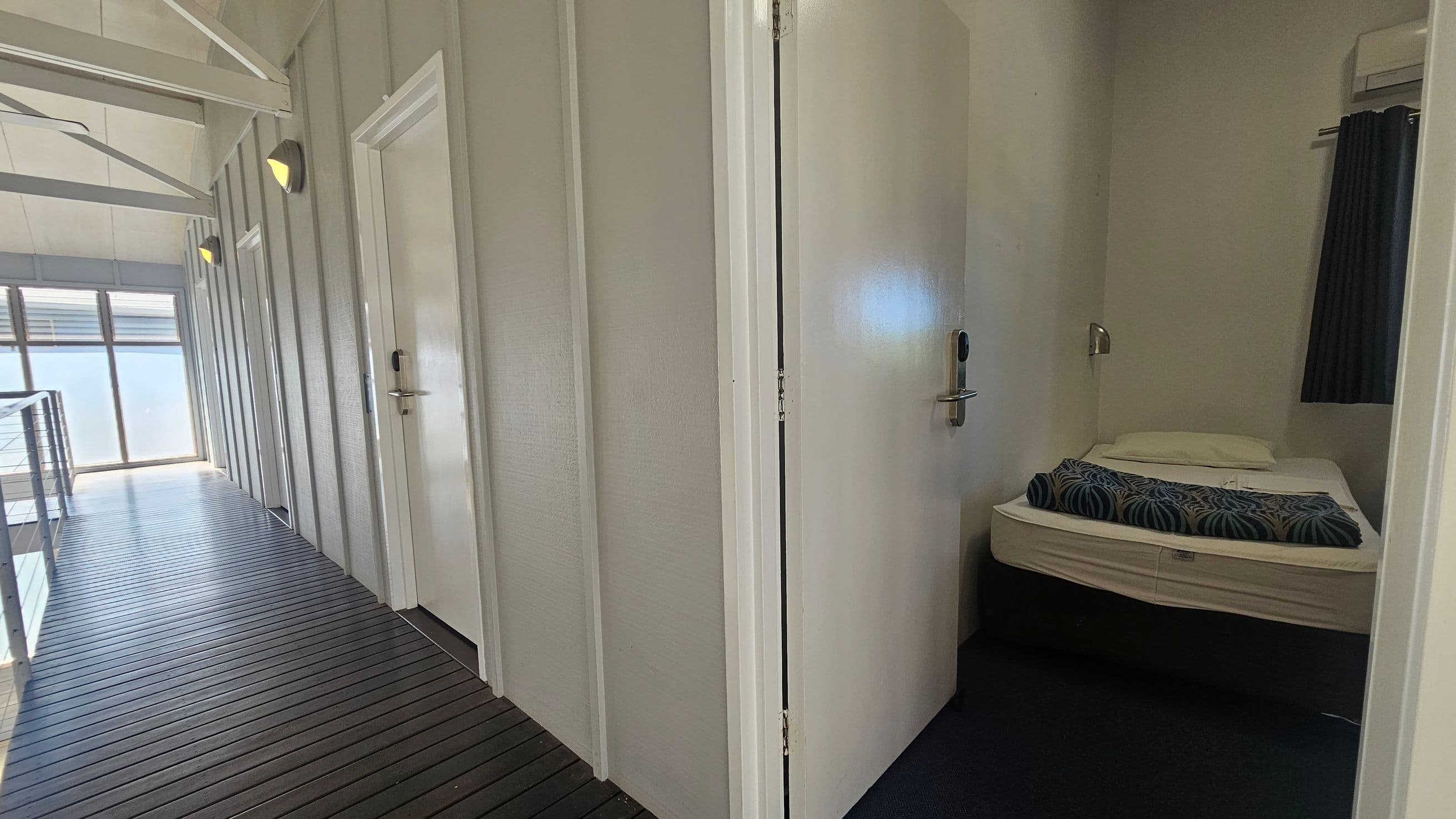 A corridor with a dark wooden floor and light-coloured walls. On the left, there are several closed doors with wall-mounted lights above them. At the end of the corridor, large windows allow natural light to enter. On the right, an open door reveals a small room with a bed, a pillow, and a patterned blanket. The room has a dark curtain and a wall-mounted light fixture. The ceiling is high with visible beams.