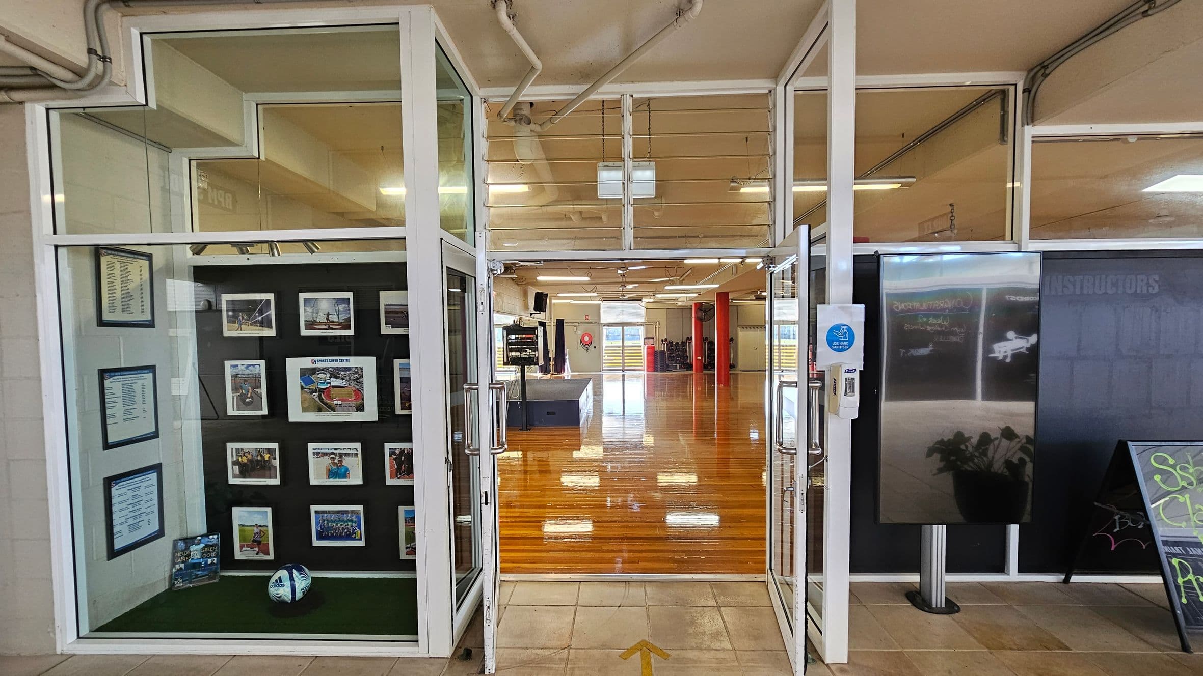 On the left, a display case with framed photos, documents, and a football on artificial grass is enclosed in glass panels within a white frame. In the middle, double glass doors with metal handles open into a room with polished wooden flooring, red columns, and white fluorescent ceiling lights. On the right, a reflective metal display screen stands next to a chalkboard sign at an angled corner. The floor in the foreground is beige tiles with an arrow pointing forward.