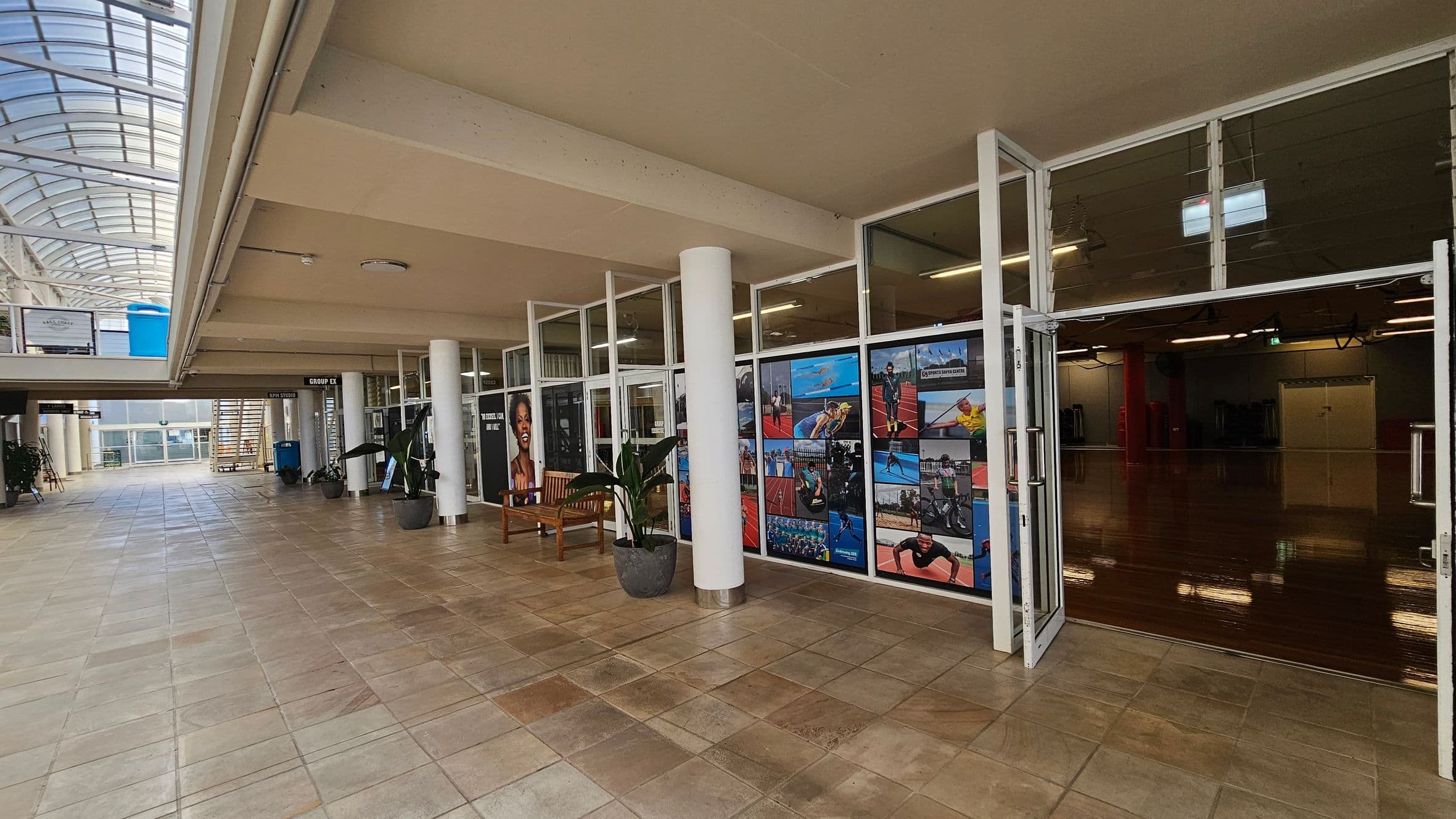 On the left, a beige tiled walkway leads to a staircase and a blue bin in the background. In the middle, white columns support a beige ceiling, with glass entrances featuring sports-themed photo panels. A wooden bench and potted plants sit in front of the glass. On the right, an open room with polished wooden flooring, red columns, and fluorescent ceiling lights. A curved glass atrium ceiling spans the left side, allowing natural light into the space.