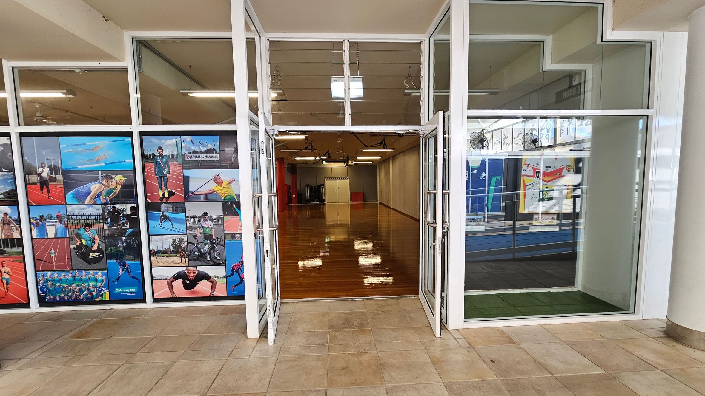 On the left, a glass wall displays several sports-themed photo panels depicting athletes in various activities. In the middle, double glass doors with metal handles open into a room with polished wooden flooring, red columns, and white fluorescent ceiling lights. On the right, a glass panel reveals an area with a blue track, posters on walls, and a section of artificial green grass. The beige tiled flooring continues across the foreground, and the ceiling is beige with white beams.