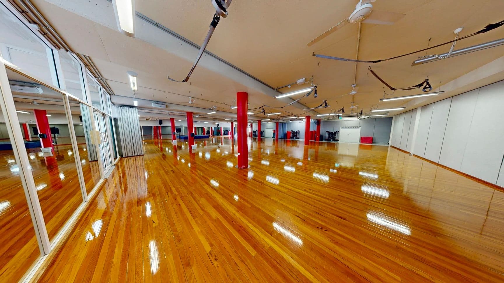 On the left, a wall of floor-to-ceiling mirrors with a section of glass panels and a visible sliding door. In the middle, a large open room with polished wooden flooring and red structural columns spaced evenly throughout. On the right, white partition walls with a small grey rear wall, some gym equipment, and a red storage cube. The beige ceiling is fitted with suspended fluorescent lights, ceiling fans, and attached fitness suspension straps.