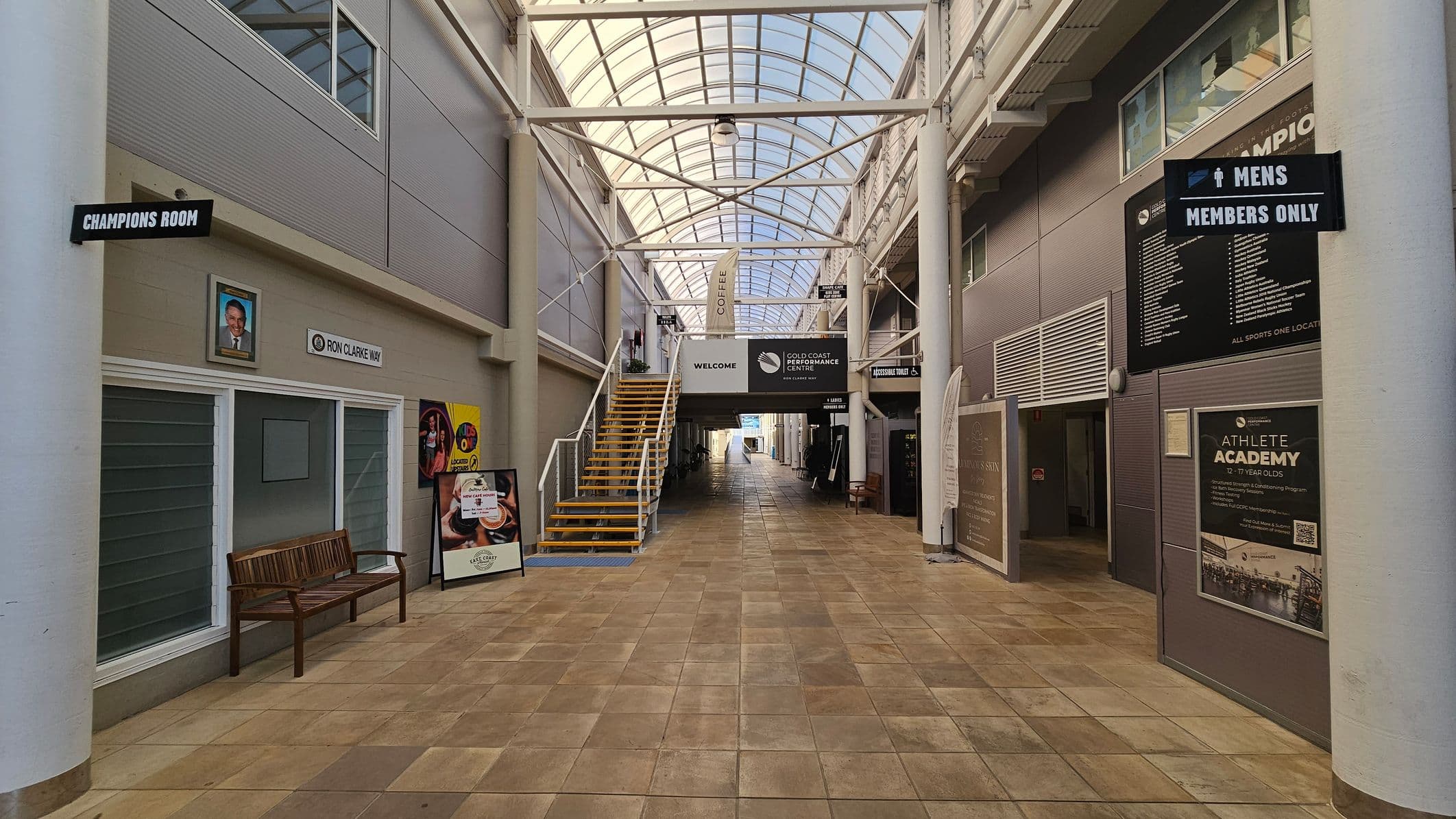 A spacious indoor corridor with a tiled floor and a high, arched glass ceiling. On the left, there's a sign for the "Champions Room" above a bench and a portrait. A staircase with yellow steps leads up to a mezzanine. On the right, signs indicate "Mens Members Only" and "Athlete Academy". Various posters and signs are displayed along the walls. The corridor extends into the distance, with natural light filtering through the ceiling.
