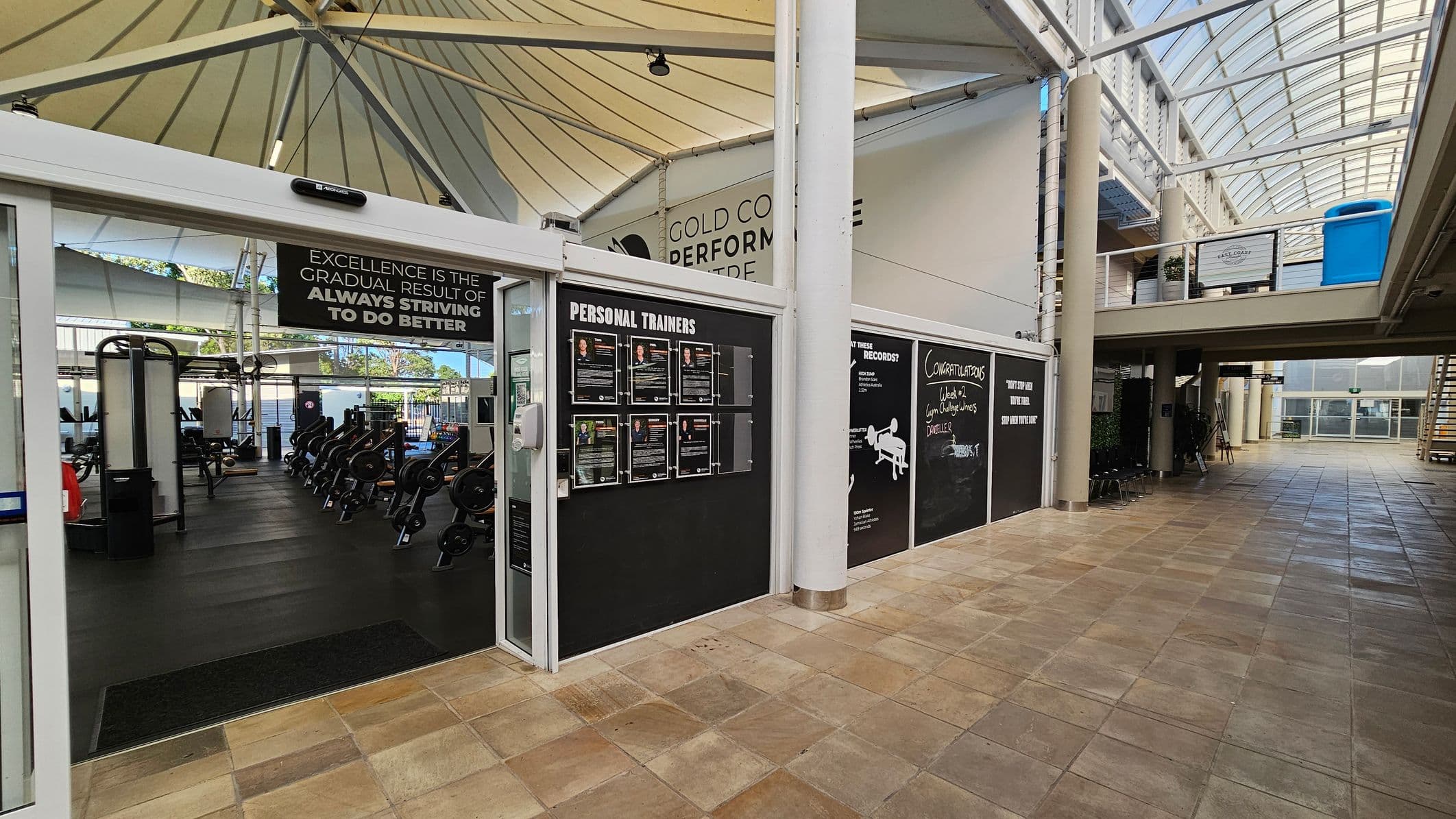 On the left, a gym entrance with exercise equipment like weights and machines visible inside. Above the entrance, a motivational sign reads, "Excellence is the gradual result of always striving to do better." In the centre, a board displays profiles of personal trainers. On the right, a corridor with tiled flooring and a glass ceiling, leading to a staircase. A congratulatory message is written on a blackboard. The ceiling is supported by white beams.