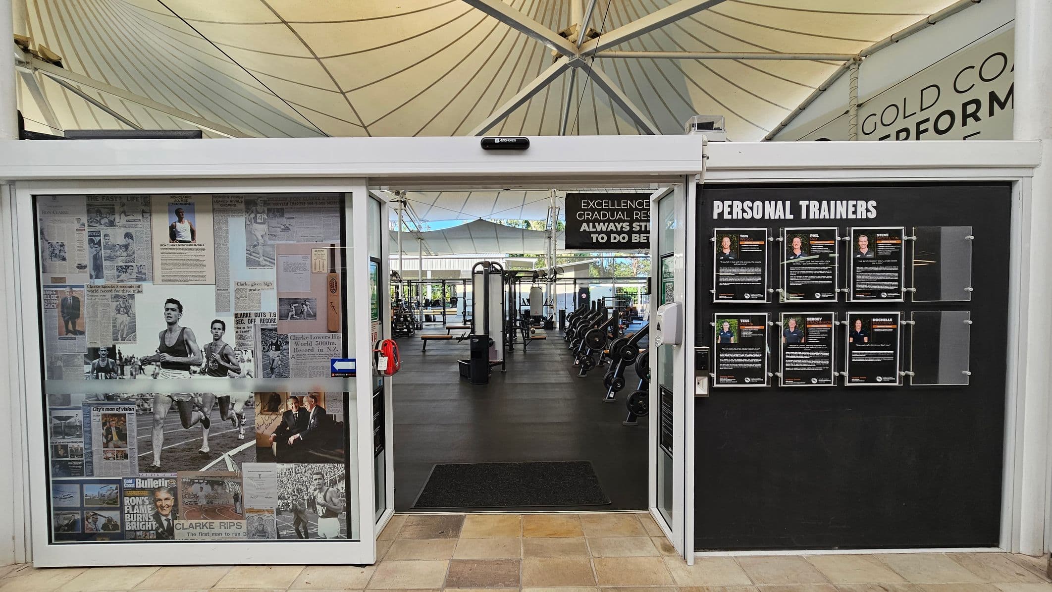 A gym entrance with a glass door on the left, covered in vintage sports newspaper clippings and photos. On the right, a black board displays profiles of personal trainers. Inside, gym equipment like weights and benches are visible. The ceiling is a white, tent-like structure with beams. The floor is tiled at the entrance and transitions to a rubber gym floor inside. A motivational sign hangs above the entrance.