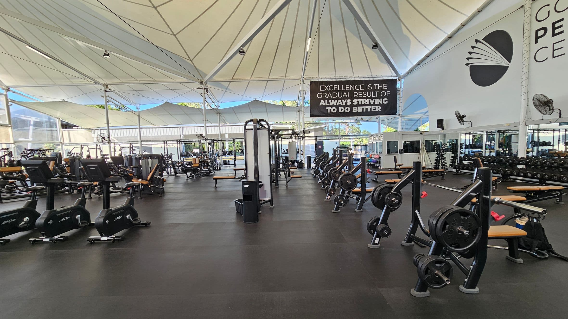 A spacious gym with a high, tent-like ceiling. On the left, there are several exercise bikes and weight machines. In the centre, a variety of weightlifting equipment is neatly arranged. On the right, a wall with a large logo and a motivational sign reads, "Excellence is the gradual result of always striving to do better." The floor is covered with black rubber mats, and large windows in the background allow natural light to fill the space.