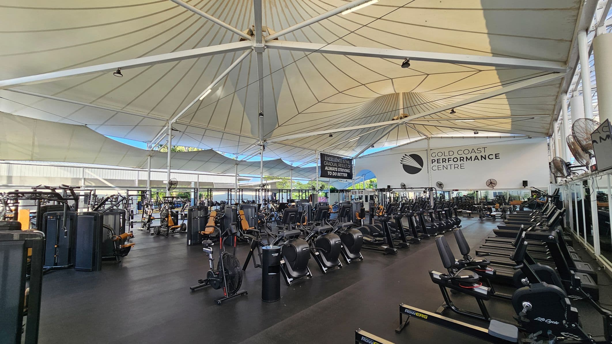 A spacious gym with a high, tent-like ceiling. On the left, there are various weight machines and exercise equipment. In the centre, rows of stationary bikes and rowing machines are neatly arranged. On the right, treadmills and additional exercise machines line the wall. The floor is dark and smooth. A large sign on the back wall reads "Gold Coast Performance Centre." The ceiling is supported by white beams, and fans are mounted on the walls.