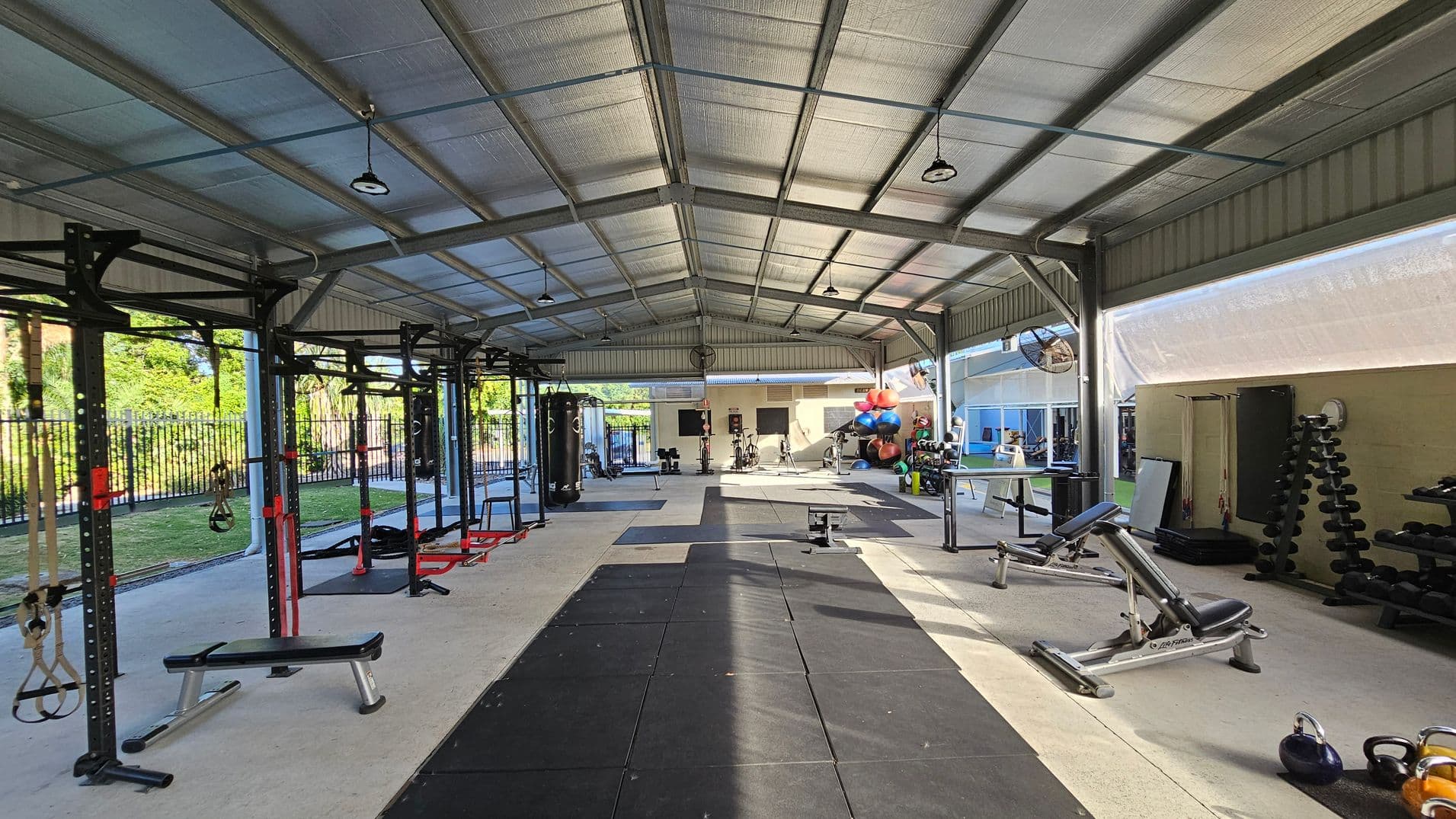 An open-air gym with a metal roof and concrete floor. On the left, there are exercise rigs with hanging straps and benches. In the centre, a black mat runs along the floor, with various gym equipment like punching bags and exercise bikes in the background. On the right, there are weight racks, benches, and kettlebells. The background shows a fenced area with greenery outside. The ceiling has exposed beams and hanging lights.