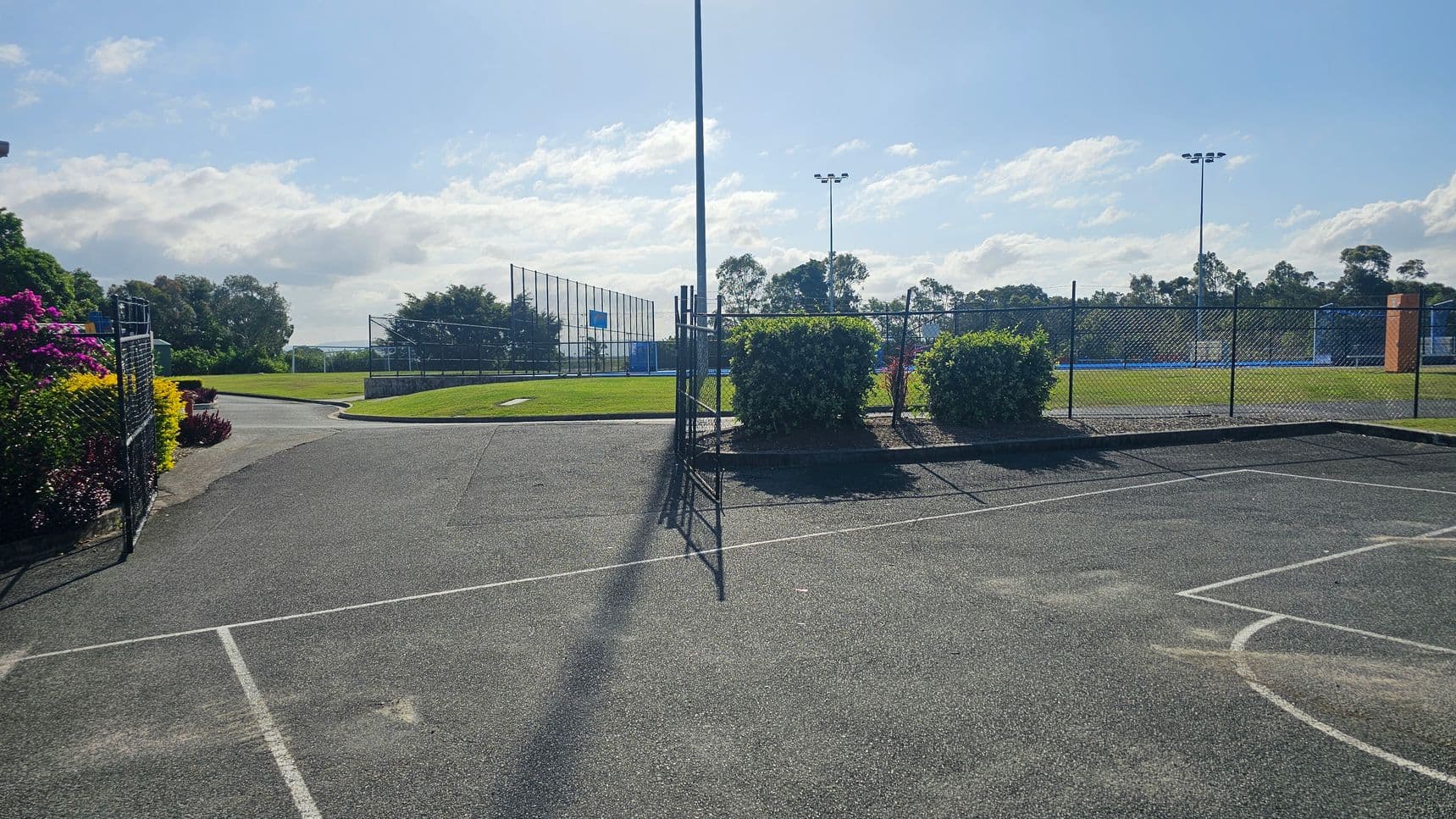 A paved sports court with white lines in the foreground. In the middle, a chain-link fence with a gate and bushes. Beyond the fence, a grassy field with a basketball court and tall floodlights. On the left, colourful flowers and a path leading to the field. Trees and a clear sky are in the background.