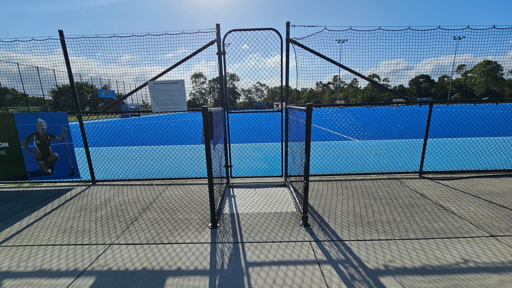 A blue sports court is visible through a black chain-link fence with a gate in the centre. On the left, there's a poster featuring a person holding a hockey stick. A sign with text is attached to the fence near the centre. The background shows trees and light poles. The ground is concrete, casting shadows from the fence.