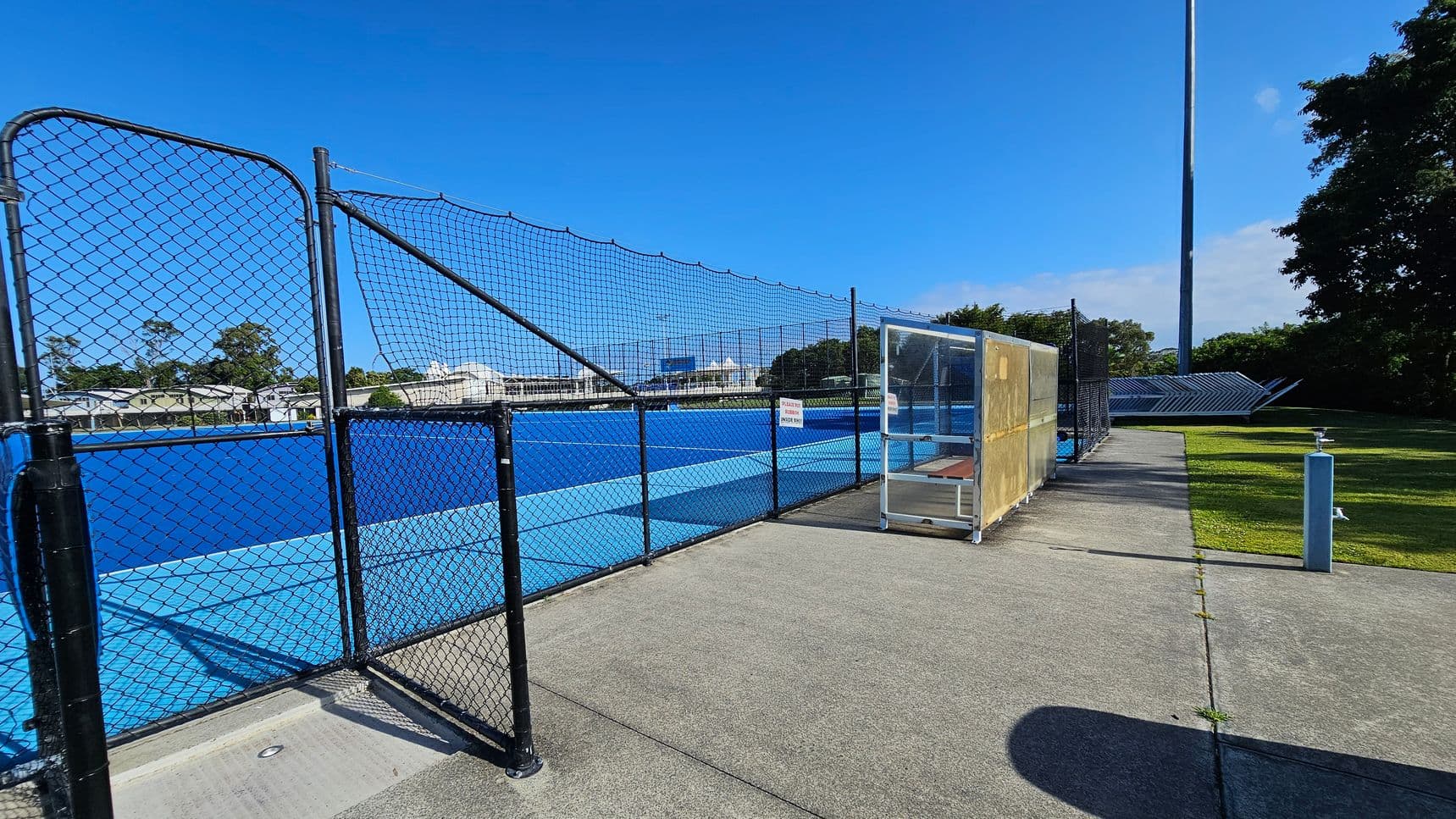 A blue sports court is enclosed by a black chain-link fence on the left. In the centre, there's a small shelter with a transparent front and a bench inside. The ground is a mix of concrete and grass, with a path leading towards the background. On the right, a tall light pole stands near some trees. A sign on the fence reads "No Public Access."