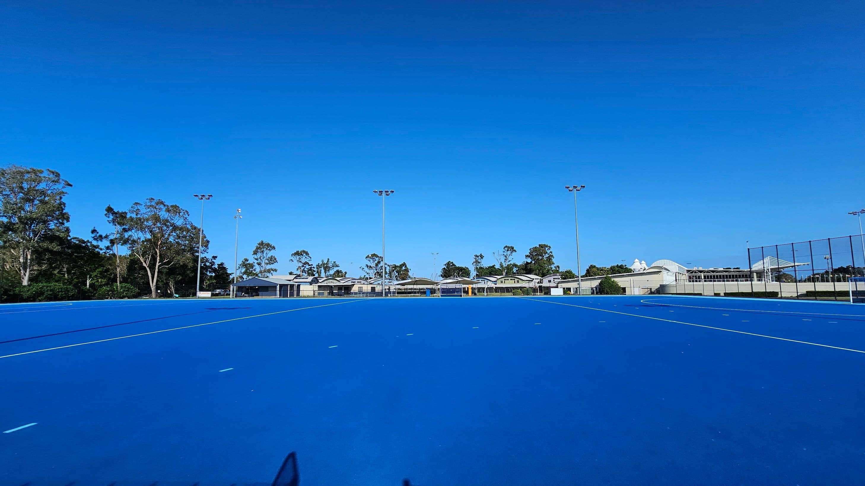 A wide blue sports field with white lines, surrounded by tall floodlights. In the background, there are several low buildings and trees. The field is enclosed by a fence on the right side. The area is open and spacious, with clear visibility of the structures and greenery in the distance.