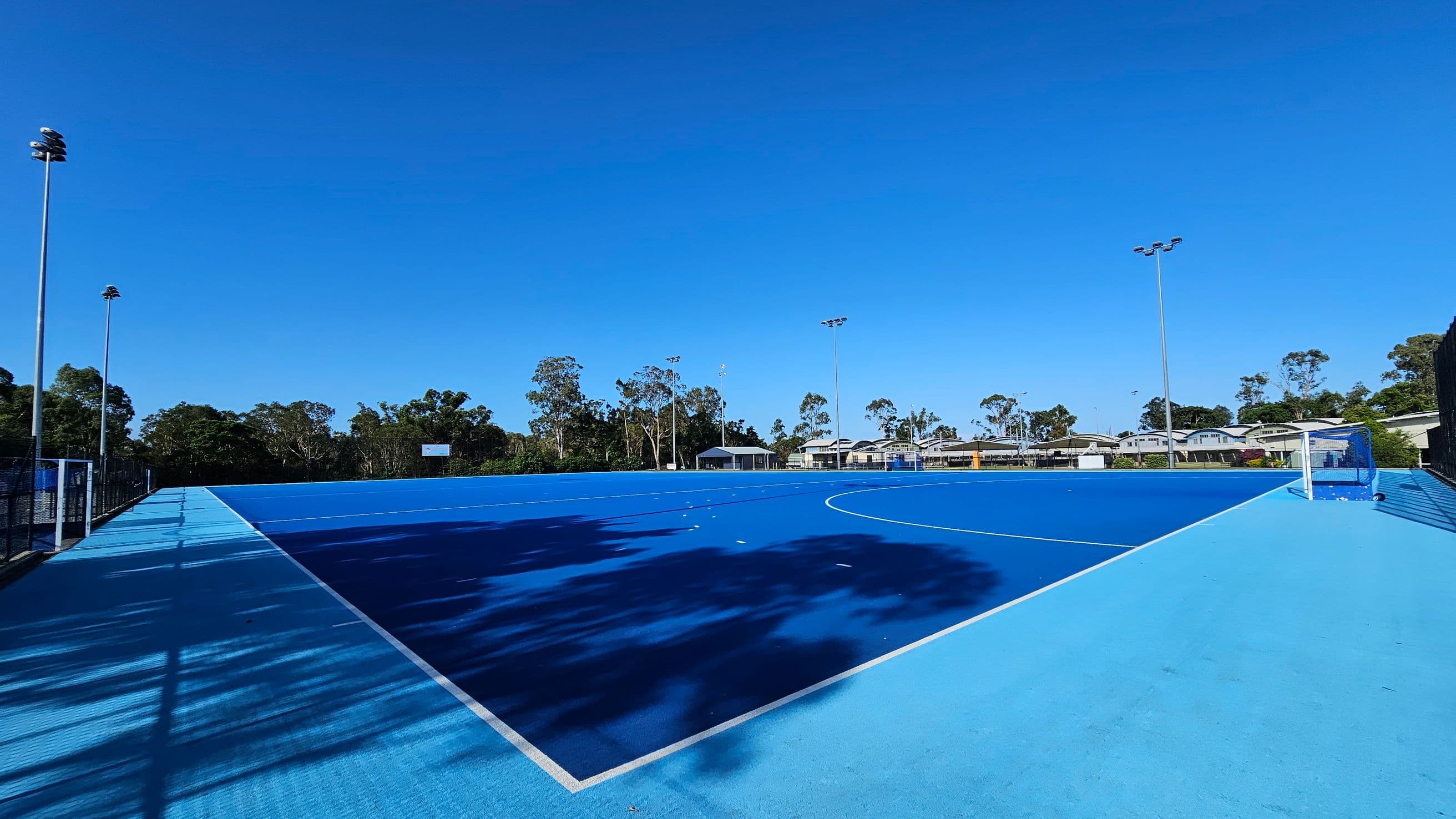 A blue sports field with white lines, possibly for hockey, is in the foreground. On the right, a goalpost is visible. Tall floodlights are positioned around the field. In the background, there are trees and a row of buildings, including a small structure with a roof. Shadows of trees are cast on the field.