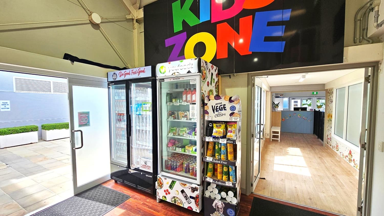 A room with a sign reading "KIDS ZONE" in colourful letters above. On the left, there are two glass-door refrigerators filled with drinks and food items. Next to them is a display stand with various snack products. The floor is wooden, and a mat is placed near the entrance. Through the open doors on the right, a hallway with light wooden flooring and decorated walls is visible, leading to a room with a small chair and a "WELCOME" sign. Outside, there are potted plants on a tiled area.