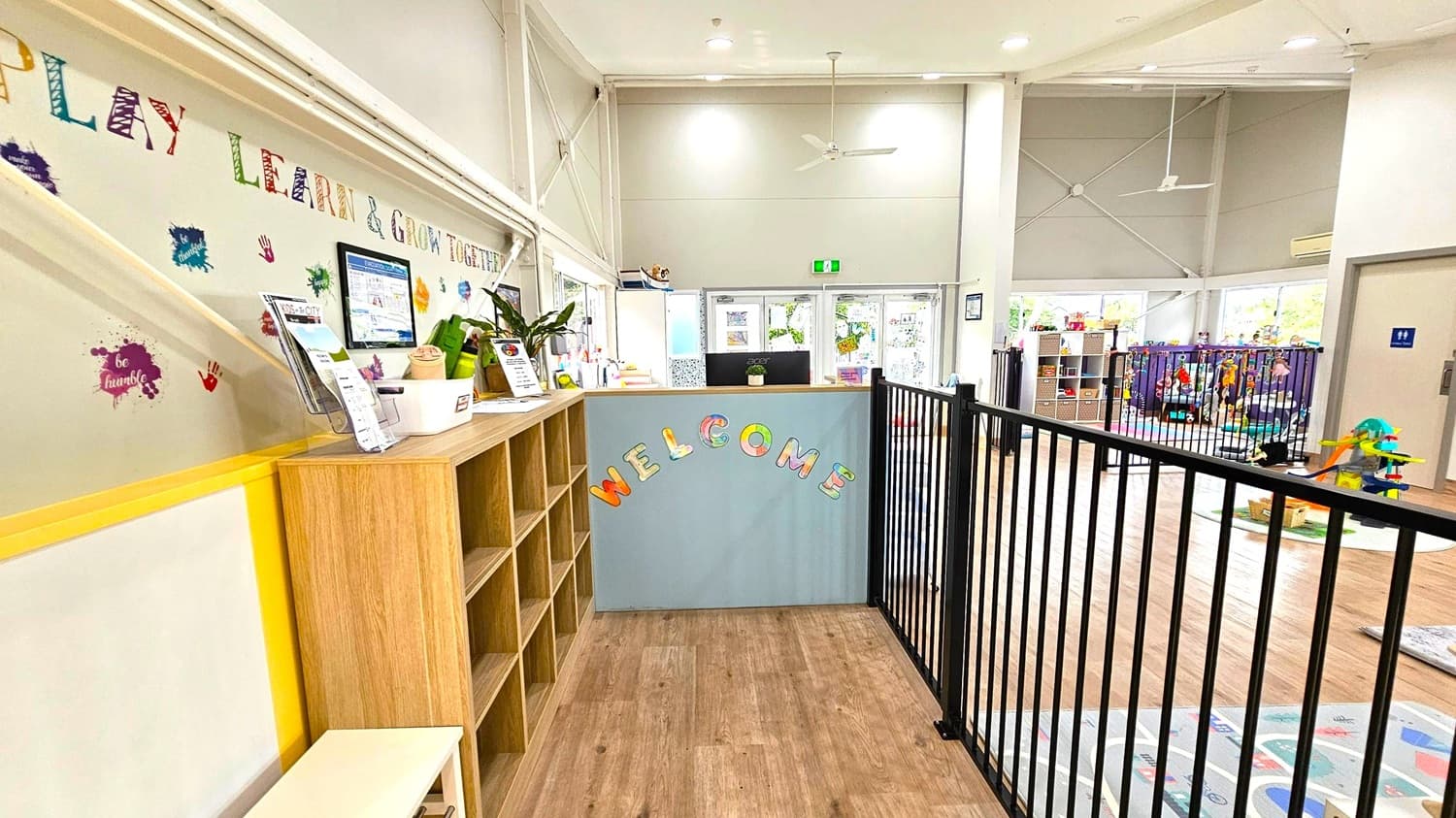 A bright, spacious room with wooden flooring. On the left, a wall displays colourful letters spelling "PLAY LEARN & GROW TOGETHER" with handprints and paint splatters. Below, a wooden shelf holds brochures and plants. In the centre, a reception desk with "WELCOME" in colourful letters. To the right, a black metal fence encloses a play area with toys and shelves. Large windows in the background let in natural light, and the ceiling is high with visible beams and a ceiling fan.