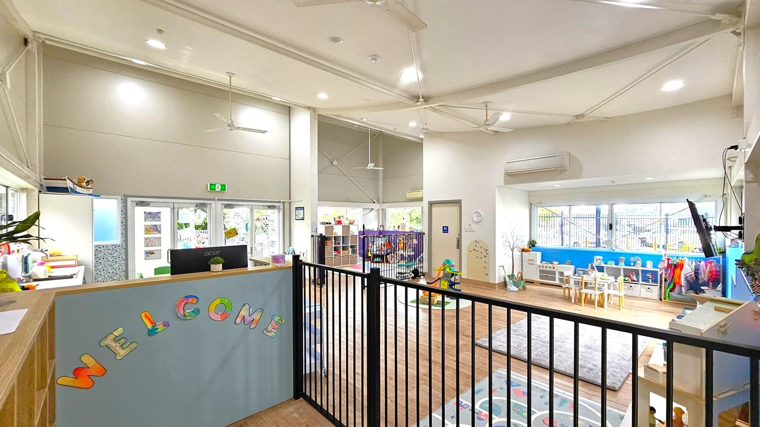 A spacious, well-lit room with a high ceiling featuring exposed beams and ceiling fans. On the left, a reception area with a "WELCOME" sign in colourful letters. In the middle, a black safety gate separates the reception from a play area with toys and a small table with chairs. On the right, a blue wall with windows, shelves, and children's coats hanging. The floor is wooden with a grey rug, and the ceiling has recessed lighting.