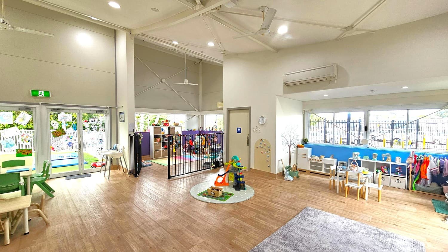 A spacious, well-lit children's playroom with wooden flooring. On the left, there are glass doors leading to an outdoor area, decorated with children's artwork. In the centre, a small play area with a toy set and a circular rug. To the right, a row of windows with a view outside, beneath which are shelves filled with toys and children's furniture. A small table and chairs are set up nearby. The ceiling is high with exposed beams and lights.