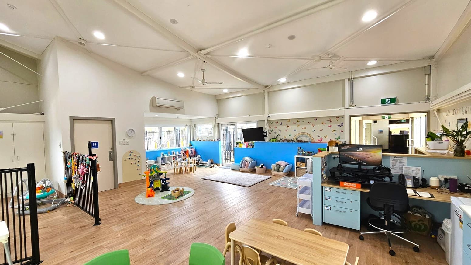 A spacious, well-lit room with a high ceiling and wooden flooring. On the left, there's a small play area with toys and a black gate. In the centre, a play area with a round rug, small tables, and chairs. The right side features a desk with a computer, chair, and office supplies. The back wall is decorated with colourful butterflies and has a TV mounted above a shelf with books. Large windows and doors allow natural light in, and there's an air conditioning unit on the wall.