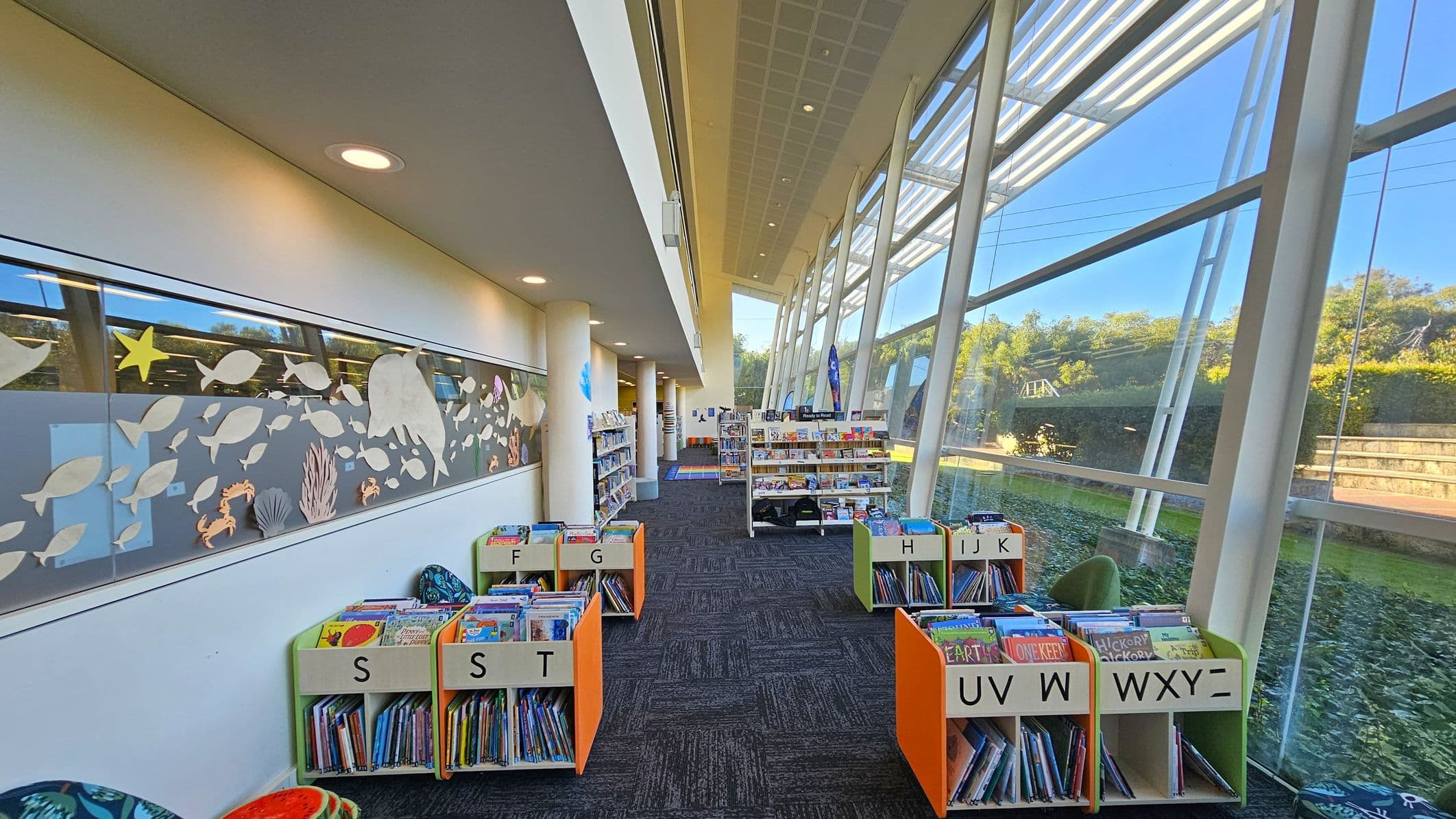 A library interior with a high ceiling and large glass windows on the right, allowing natural light to illuminate the space. On the left, a wall features a decorative panel with cut-out shapes of fish and sea creatures. The floor is carpeted in a dark pattern. In the centre, there are low bookshelves with books organised alphabetically, labelled with letters. Further back, taller bookshelves are visible, and outside the windows, greenery and a pathway can be seen.