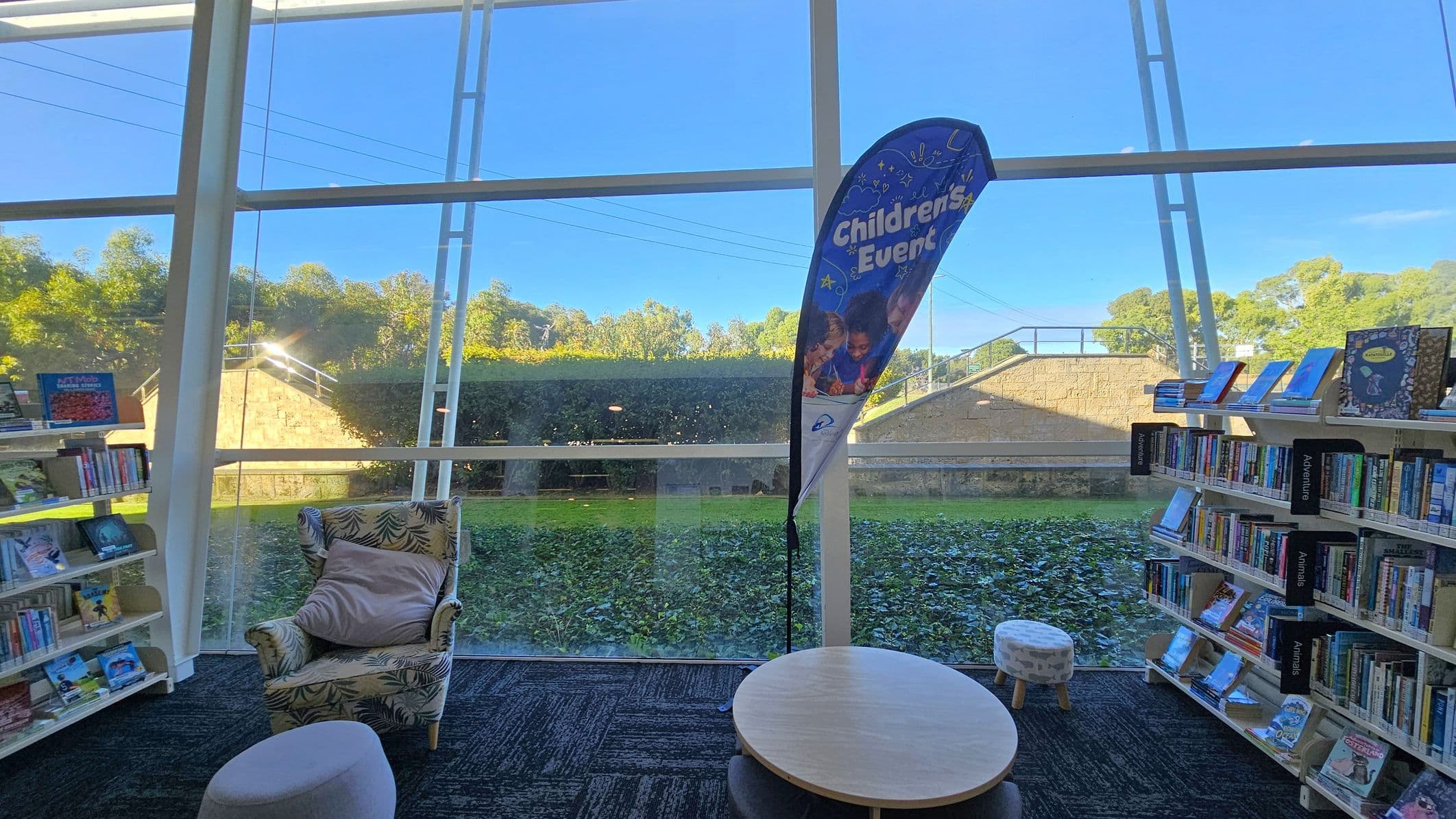 A library interior with large glass windows in the background, revealing a view of greenery and a stone wall outside. In the foreground, there is a patterned armchair with a cushion, a small round table, and a stool. To the left and right are bookshelves filled with books, with signs indicating sections like "Adventure" and "Animals." A banner stands near the window, advertising "Children's Events." The floor is carpeted in a dark pattern.