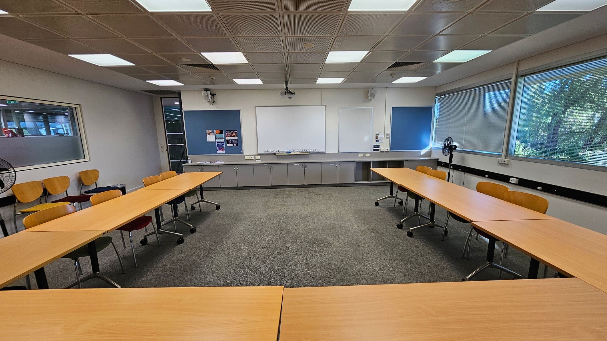A classroom with a U-shaped arrangement of wooden tables and chairs on a grey carpeted floor. The back wall features a large whiteboard flanked by blue noticeboards with some posters. Below the boards are grey cabinets. On the right, large windows with blinds allow natural light in, and a fan is positioned nearby. The ceiling has a grid pattern with fluorescent lights. On the left, a glass panel separates this room from another area.