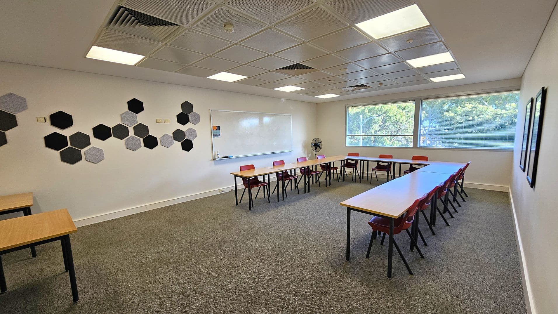 A classroom with a U-shaped arrangement of tables and red chairs. On the left wall, there are hexagonal acoustic panels in various shades of grey and black. The back wall features a large whiteboard. The right side has large windows with blinds, allowing natural light to enter. The ceiling is fitted with square lights and ventilation panels. The floor is carpeted in grey.
