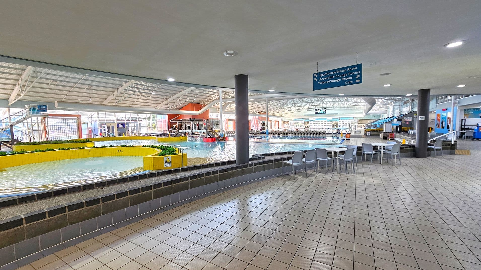 An indoor swimming pool area with a tiled floor. On the left, there's a shallow circular pool with yellow tiled edges and some plants. In the centre, a larger pool with clear water is visible, surrounded by a spacious area with tables and chairs. The ceiling is high with metal beams and lights. On the right, there are more seating areas and signs indicating directions to facilities like a spa, sauna, and café. The background shows large windows and additional pool features.