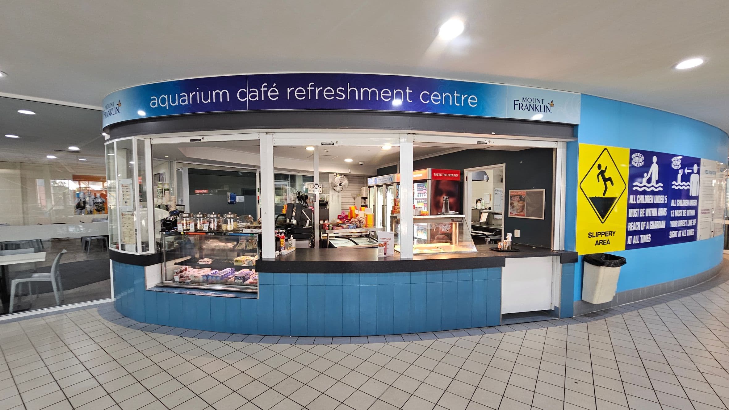 Aquarium café refreshment centre with a curved counter displaying snacks and drinks. The counter is blue with a black top. Behind the counter, there are shelves with various food items and a drinks fridge. On the right, a yellow "Slippery Area" sign and blue safety notices are visible. The floor is tiled in a light colour, and the ceiling has recessed lighting. To the left, there are tables and chairs in a seating area.