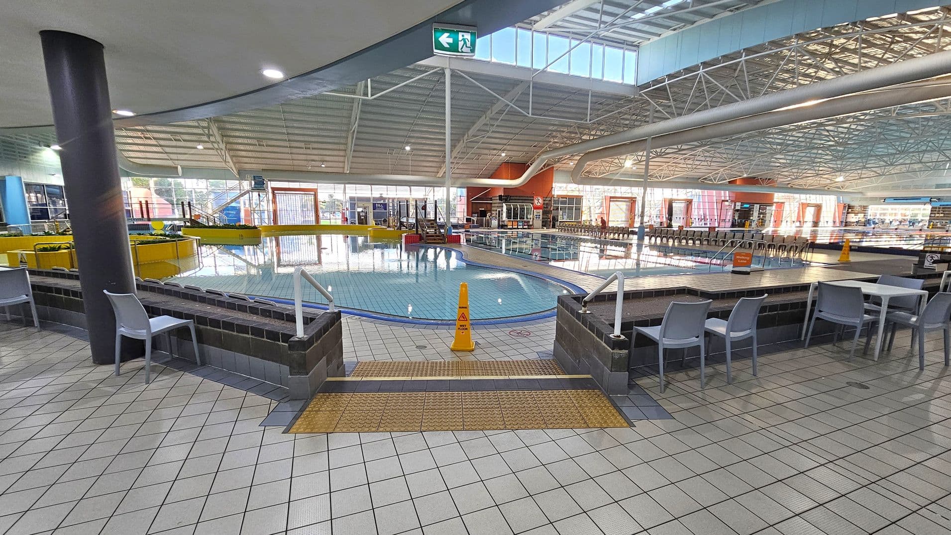Indoor swimming pool area with a tiled floor. In the foreground, there are tables and chairs on either side. A small pool with a yellow safety cone is in the centre, surrounded by a low brick wall. The background features a larger pool with a slide and a row of chairs along the far wall. The ceiling is high with metal beams and large windows allowing natural light. An exit sign is visible above.